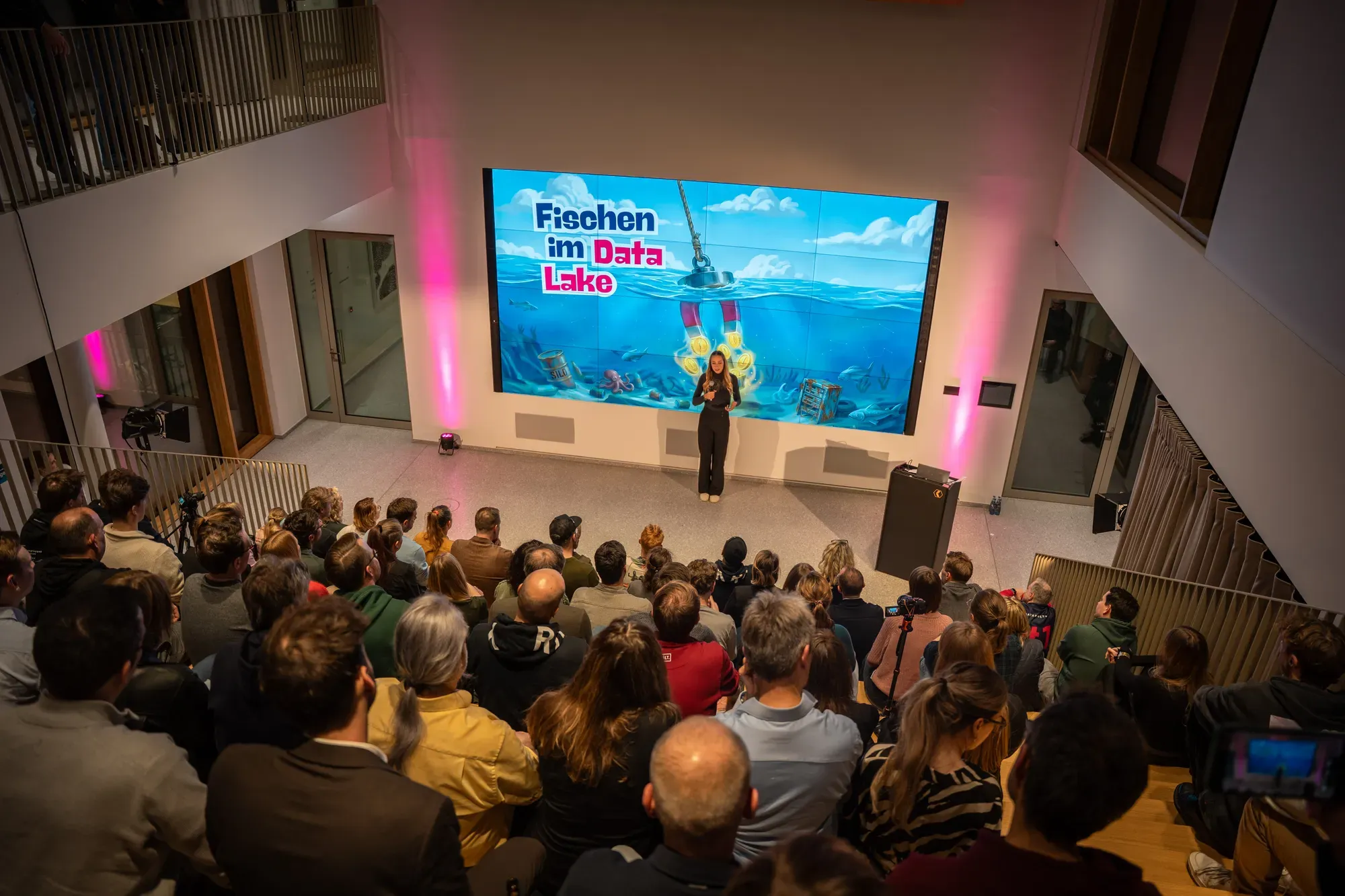 Publikum im Atrium beim DSLAM Münster 2026, Alex Ulbricht vor der großen Leinwand